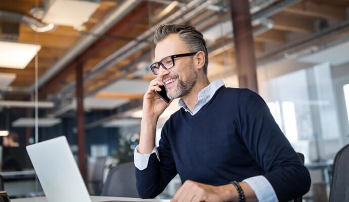 Hombre que sonríe con el celular en la oreja mientras mira una computadora portátil